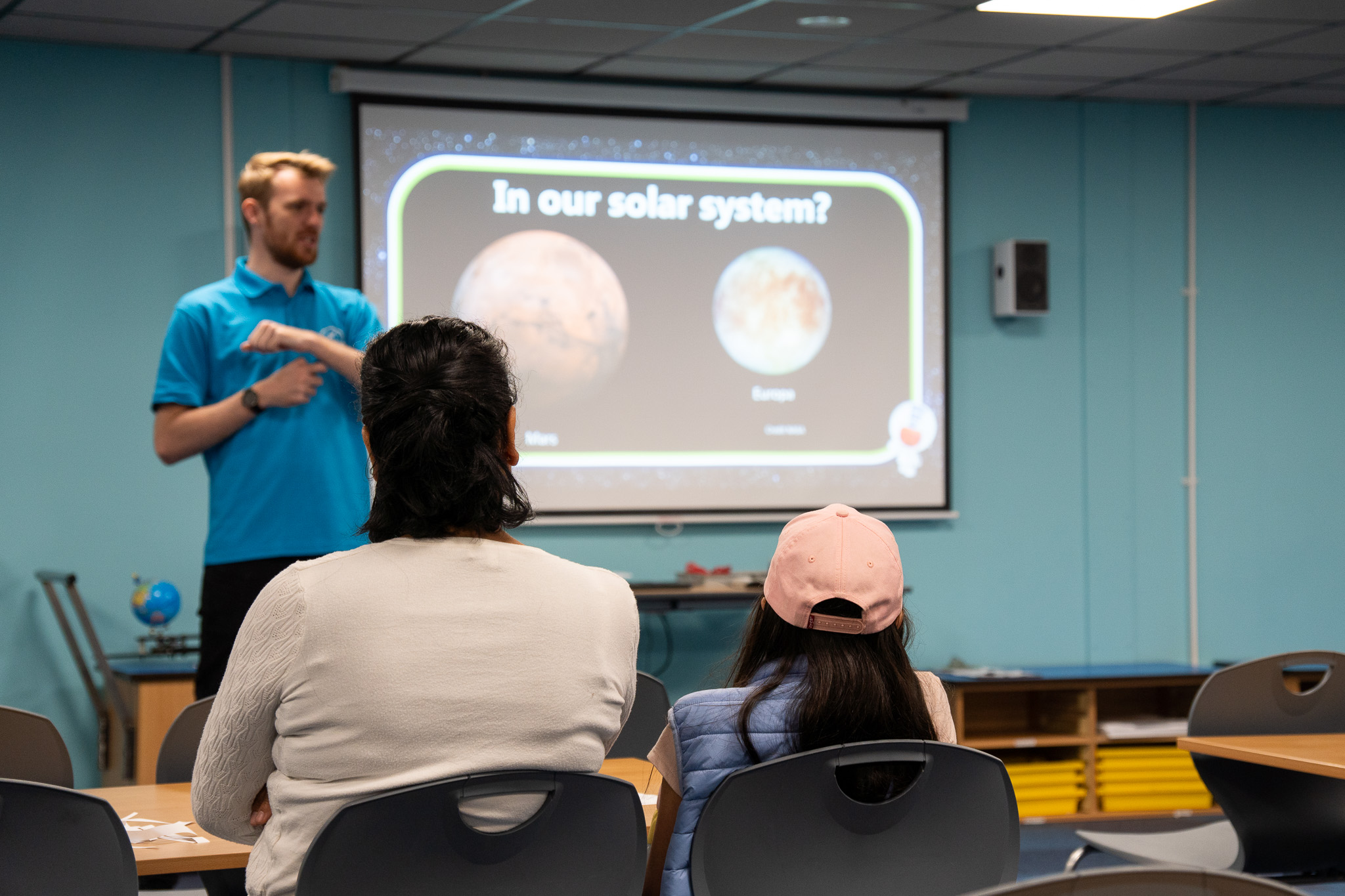 A presenter from the Education team presenting a PowerPoint to a classroom in the background, with a child and their parent shown in the foreground, listening.