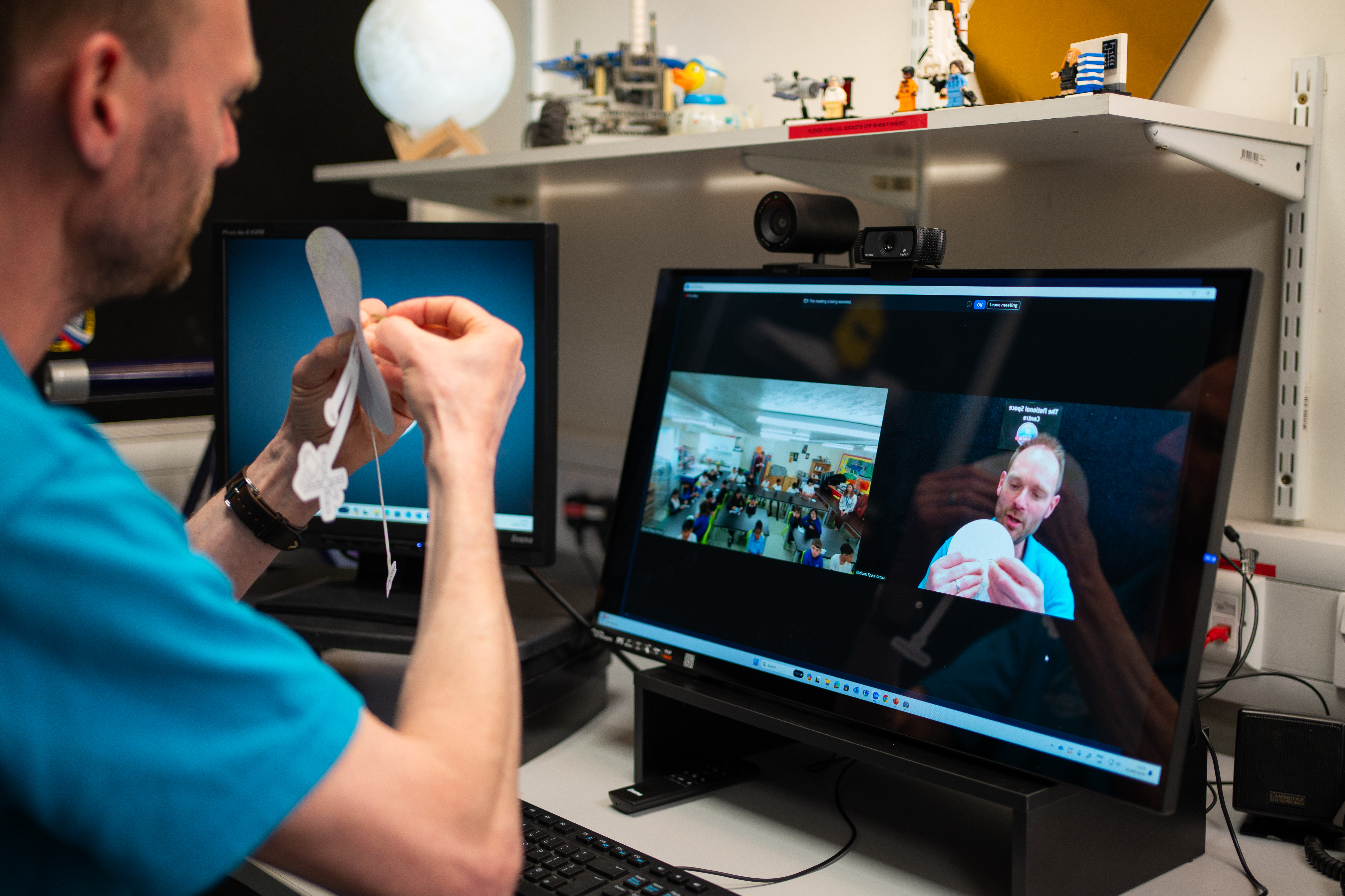 An Education Presenter holding a paper earth with paper satellites attached, presenting to a school group on a Video Conference.