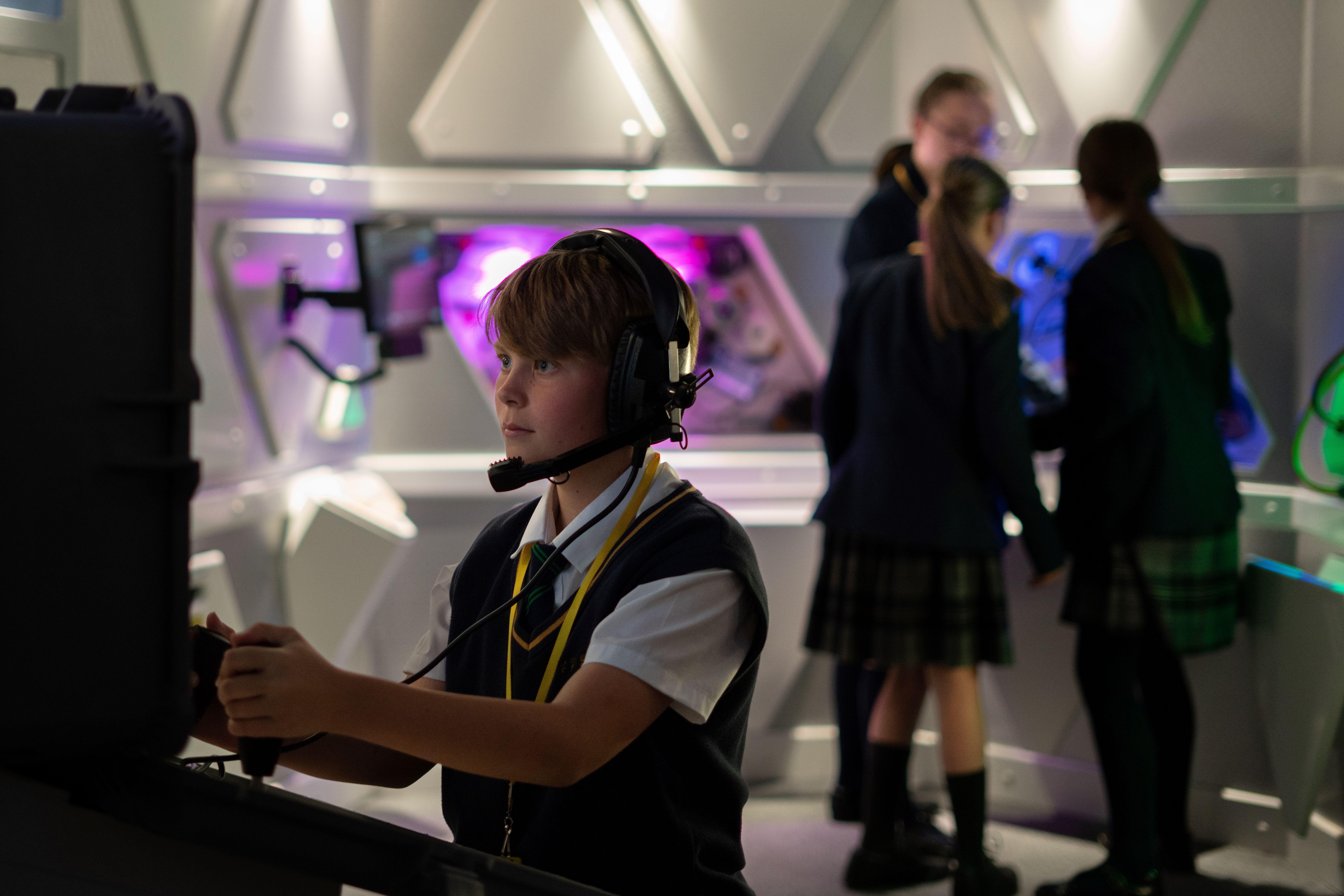 A child with headphones and a microphone on a computer controlling a Mars Rover. Other children are talking in the background.