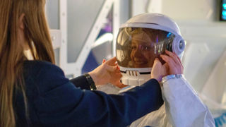 Two school children, one dressed in an astronaut costume, with the other helping adjust their helmet, stood in a Mars base.