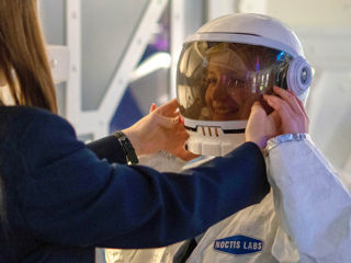 Two school children, one dressed in an astronaut costume, with the other helping adjust their helmet, stood in a Mars base.
