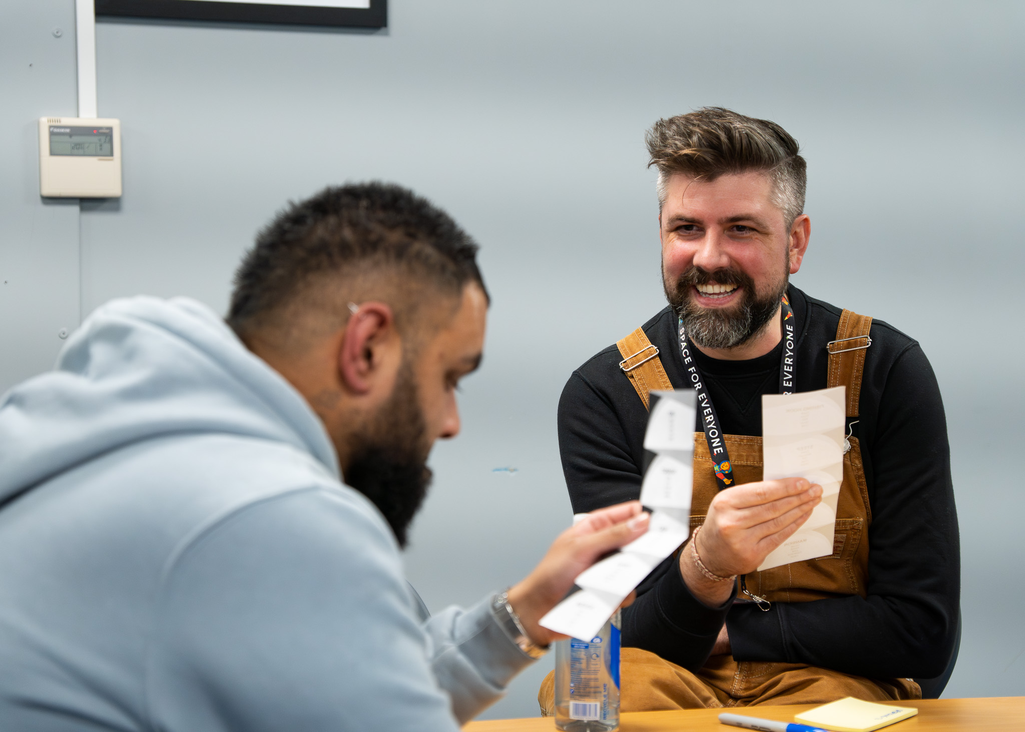 One person is reading off a small piece of paper, another person looks at them. They are both smiling.