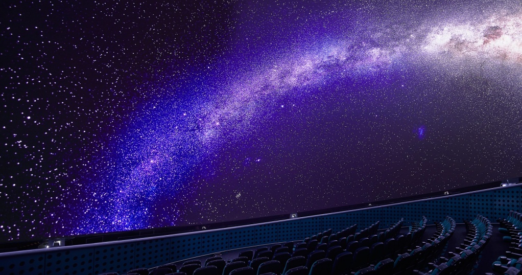 Planetarium seating is seen at the bottom of the image. The planetarium screen displays an image of the milky way galaxy as visible from Earth.